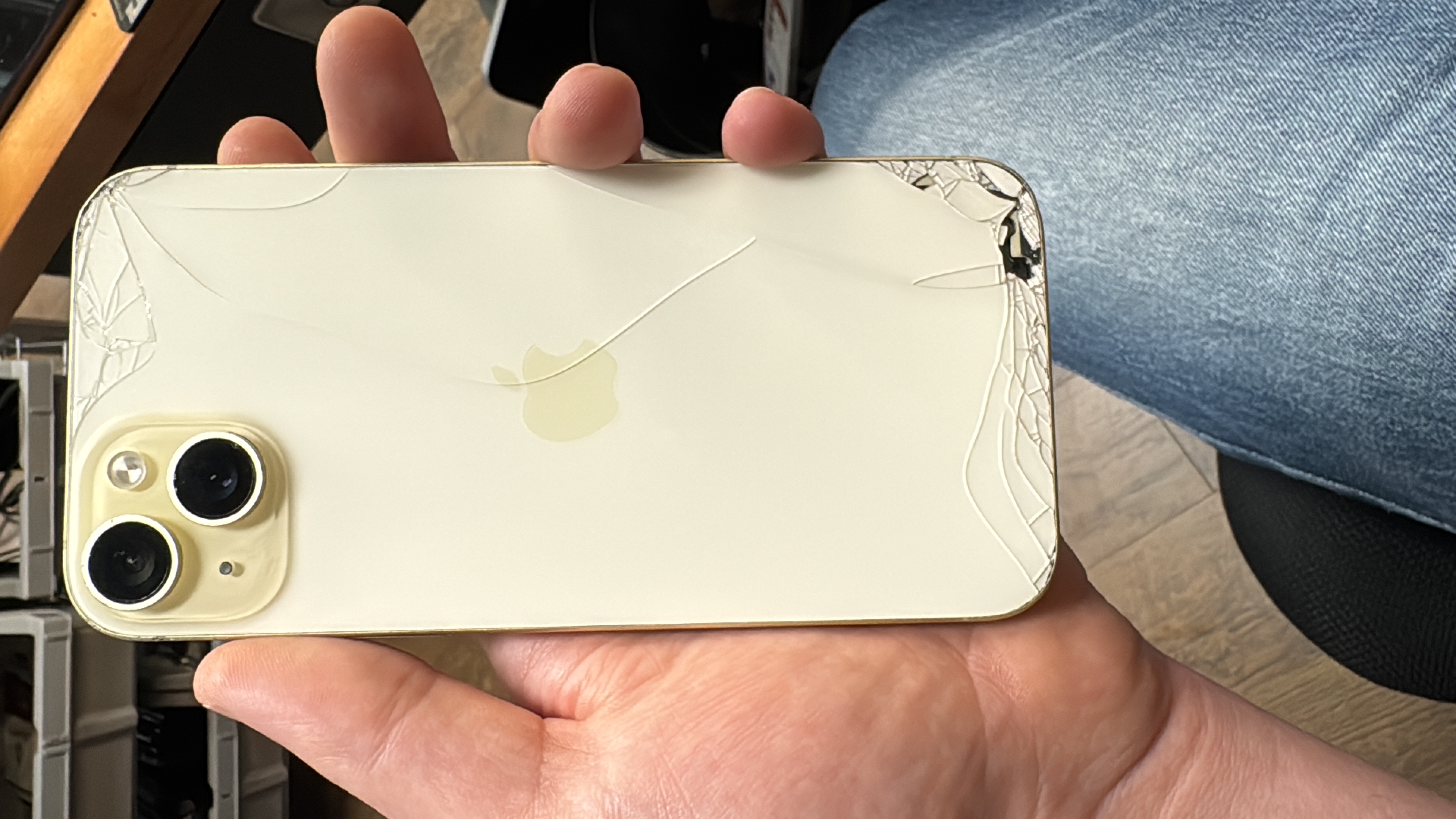 Cracked back glass on a yellow iPhone 15 held at the Charlotte Tech Repair bench, with cracking running from the lower corner