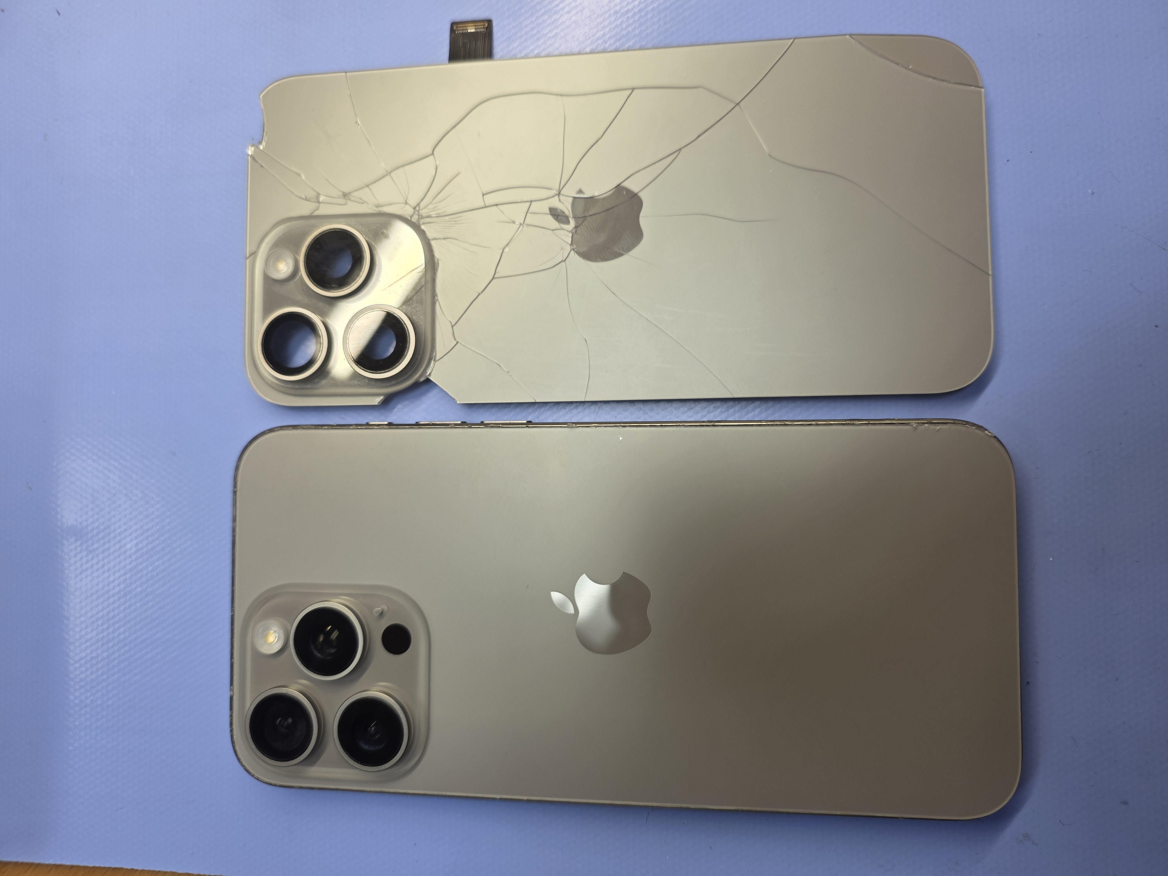 A repaired iPhone 15 Pro with new back glass beside the cracked panel that was removed from it