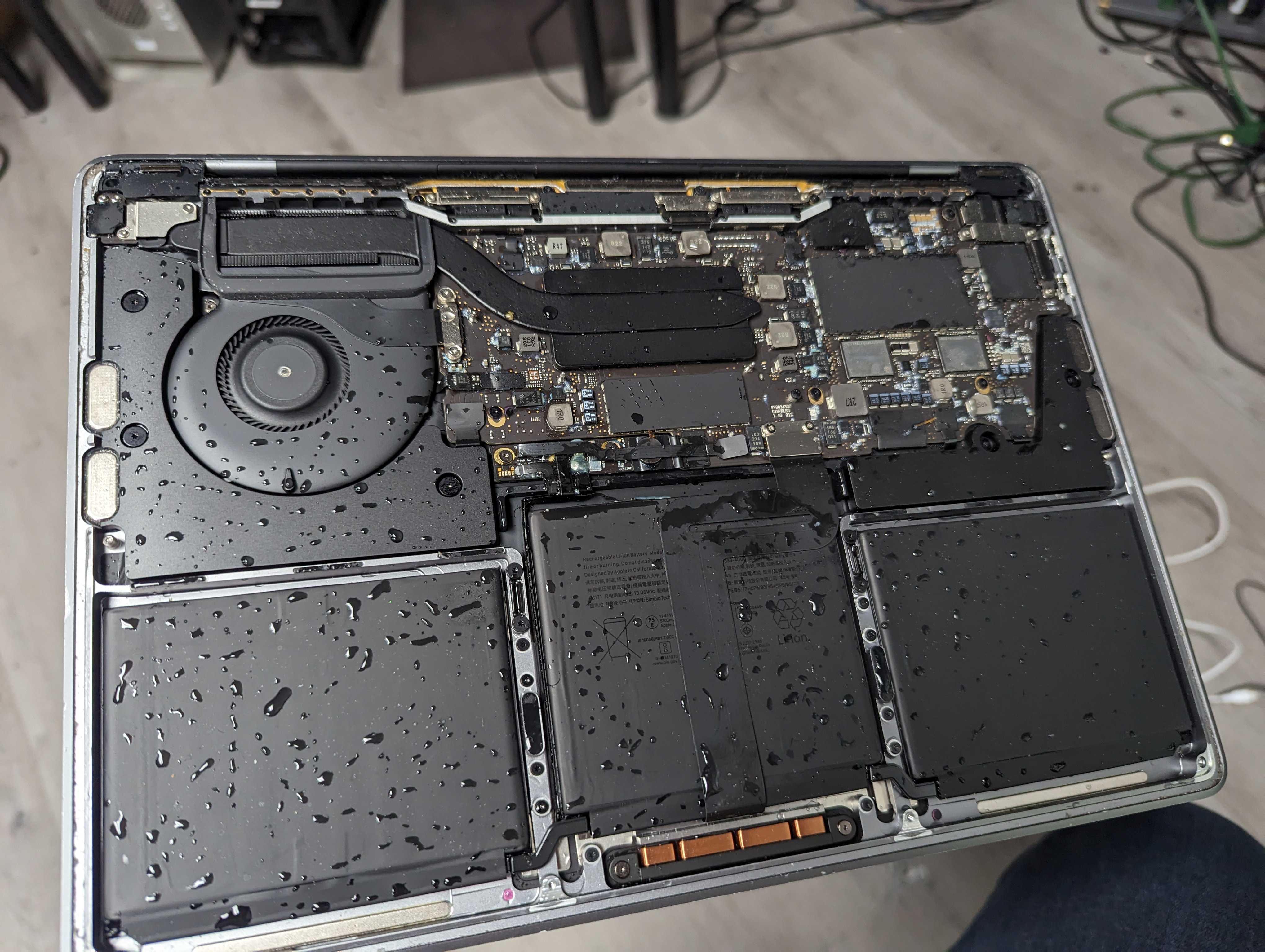 A MacBook Pro opened immediately after a liquid spill, with water droplets covering the logic board, battery, and interior case