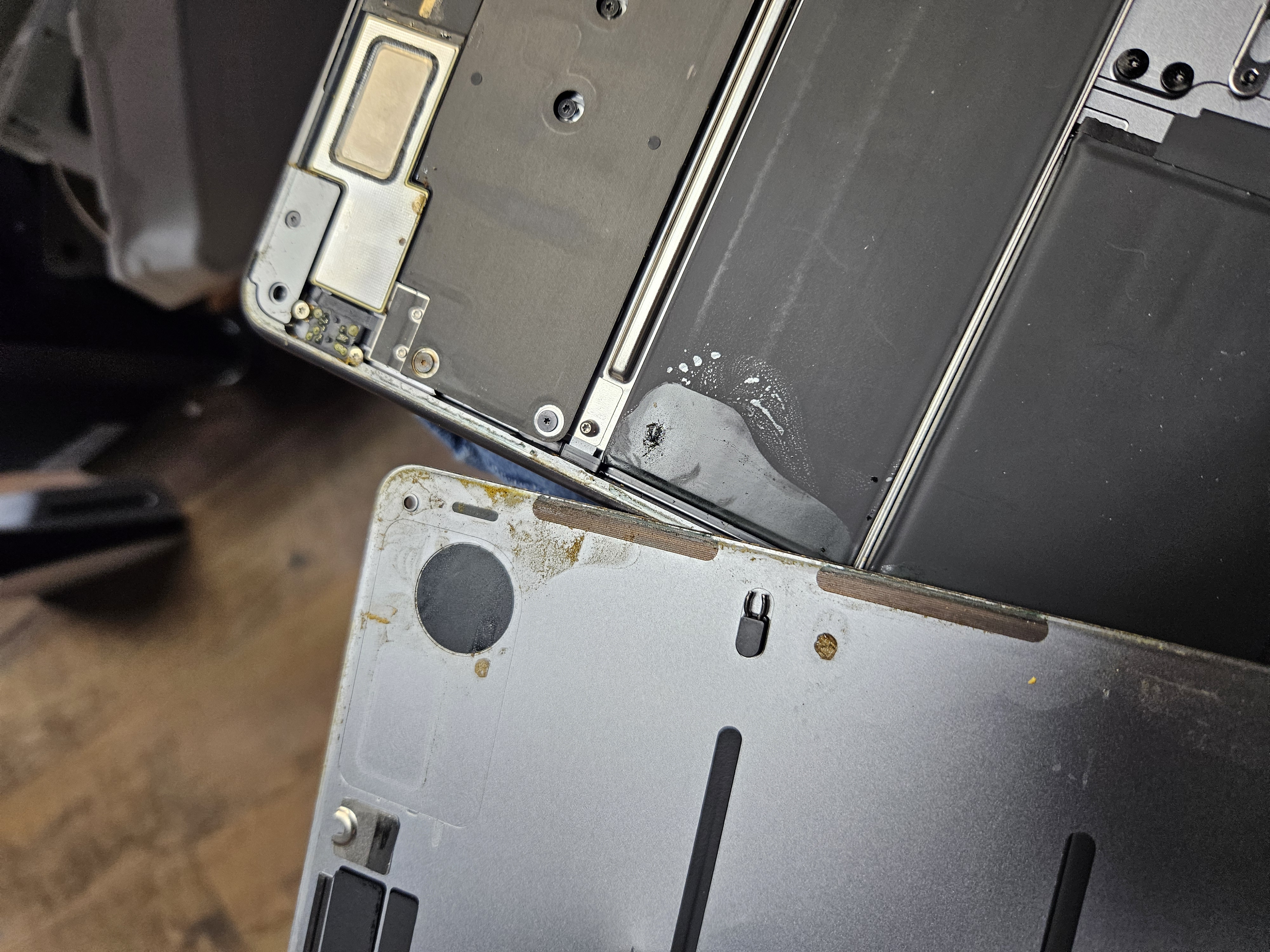 Liquid damage inside a MacBook with the bottom case removed, showing corrosion spreading across the frame and residue on the battery
